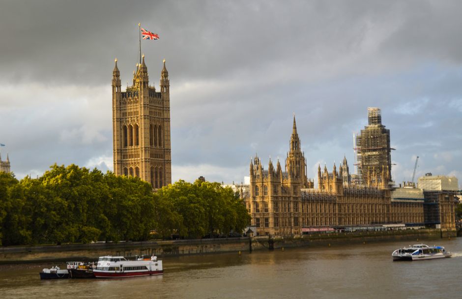 A daytime view of the Palace of Westminster in Whitehall Parliament Square, Westminster, with the iconic Elizabeth Tower and Big Ben clock tower clearly visible against a bright blue sky. The Gothic-s